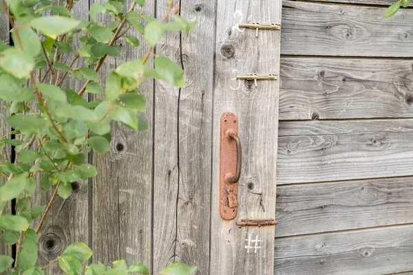 Drzwi starej drewnianej szopy lub toalety z zardzewiałą klamką i trzema zatrzaskami, obok drzewa, we wsi. — Zdjęcie stockowe