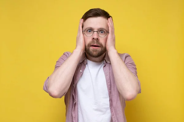 Caucasian man with glasses is surprised and bewildered, he presses hands to head and looks anxiously with large eyes. Yellow background. — Stock Photo, Image