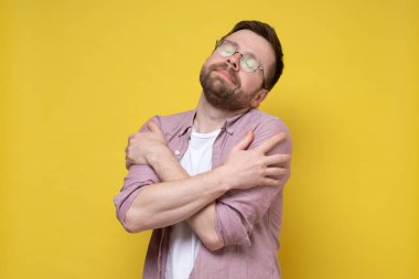 Sevimli, sakallı, gözlüklü adam nazikçe kendine sarılıyor ve rüyasında gözlerini kapıyor. Sarı arkaplan.