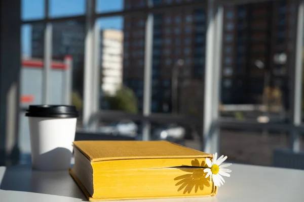 Geschlossenes gelbes Buch mit Lesezeichen mit Kamillenblüte und aromatischem Kaffee in einem Einwegbecher aus Papier auf dem Tisch in einem Café. — Stockfoto