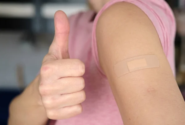 Zbliżenie żeńskiego ramienia z bandażem po szczepieniu przeciw kovidowi-19 i gest zatwierdzenia, kciuk w górę. Koncepcja zdrowia. — Zdjęcie stockowe