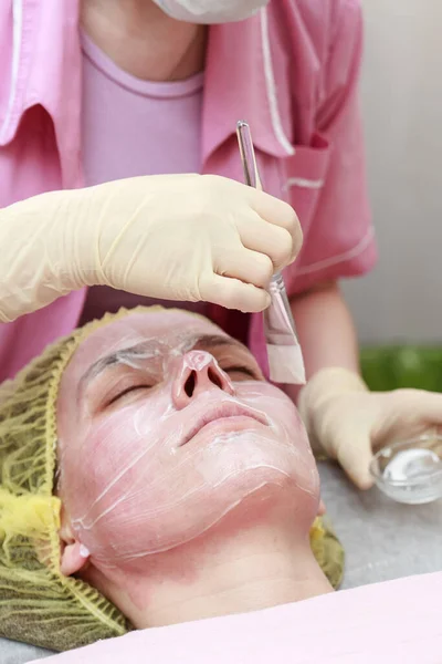 Kosmetolog använder en borste för att applicera en lugnande, vit mask på patientens rodnade hud, efter kemisk peeling. — Stockfoto
