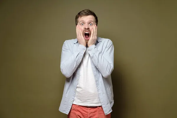 Verängstigter, schockierter Mann hält Gesicht mit Händen und schreit mit weit aufgerissenen Augen. Grüner Hintergrund. — Stockfoto