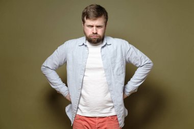 Beyaz sakallı adam kaşlarını çattı, bir şeyden memnun değil ve elleri kalçasında kızgın görünüyor. Yeşil arkaplan.