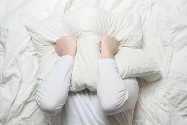 Muž v bílém pyžamu nemůže kvůli hluku spát a ve stresu přikrývá hlavu polštářem. Koncept nespavosti. Horní pohled. — Stock fotografie