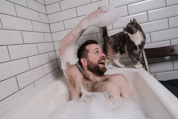 Satisfeito, homem sorridente banha-se em um banho com espuma exuberante quando seu gato amado se aproximou dele e olha com interesse. — Fotografia de Stock