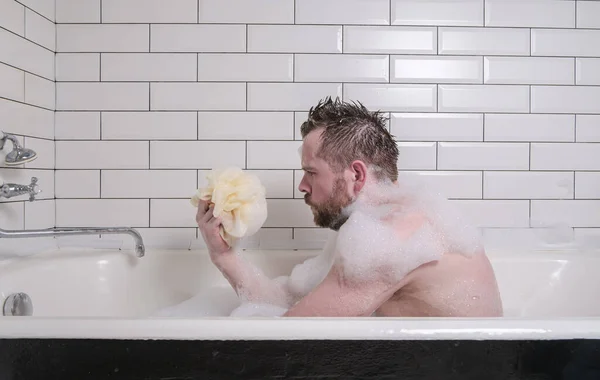 Smutny człowiek kąpie się w wannie z bujnymi piankami mydlanymi, trzyma gąbkę w dłoniach i troskliwie patrzy. — Zdjęcie stockowe