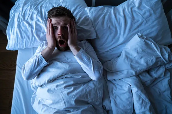 El hombre tuvo una pesadilla terrible, se despertó horrorizado, mirando asustado con grandes ojos a la cámara. Insomnio y concepto de salud mental. — Foto de Stock