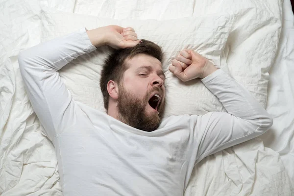 Cute bearded man wakes up from sleep, he yawns and stretches as he lies in bed. Top view. Close-up. — Stock Photo, Image