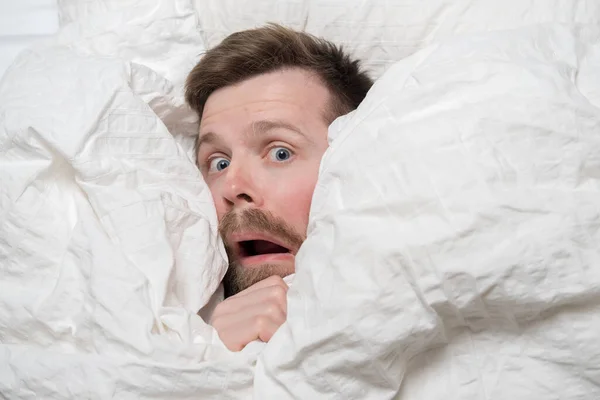 Caucasian man woke up from a nightmare, he looks out from under the blanket in fright and looks with big eyes. Top view. — Stock Photo, Image