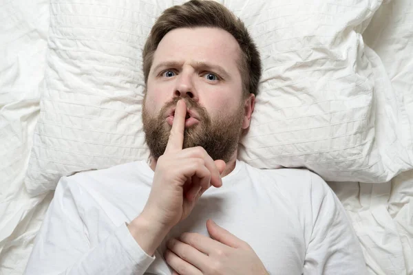 El hombre serio yace en la cama y pide guardar silencio con el dedo en los labios. Silencio o concepto secreto. Vista superior. Primer plano. — Foto de Stock