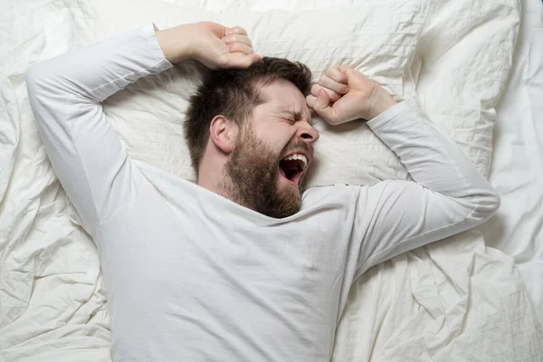 El hombre barbudo en pijama blanco está cansado y quiere dormir, bosteza y se estira mientras está acostado en la cama. Vista superior. Primer plano. — Foto de Stock