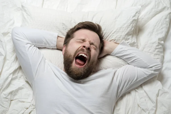 Cute bearded man wakes up from sleep, he yawns and stretches as he lies in bed. Top view. Close-up. — Stock Photo, Image