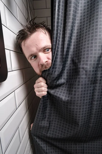 Alarmed, wet man looks out of the shower, bites the curtain with excitement and looks away in dismay. — Stock Photo, Image