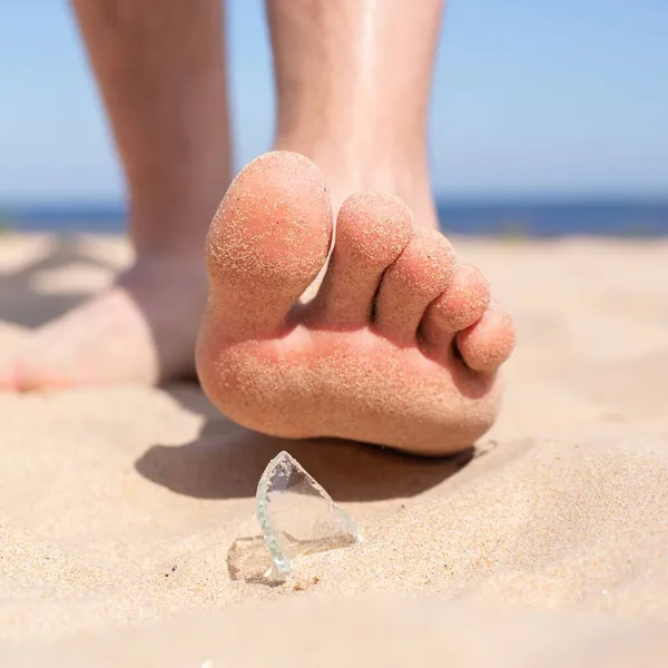 Leg walks on the beach and runs the risk of injury, stepping on a shard of broken glass, which is lying on the sandy seashore. — Stock Photo, Image