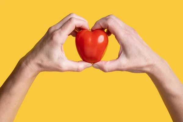 Red tomato in the form of a heart, a symbol of love in hands, on a yellow background. Healthy food concept. Valentines Day. Top view. — Stock Photo, Image