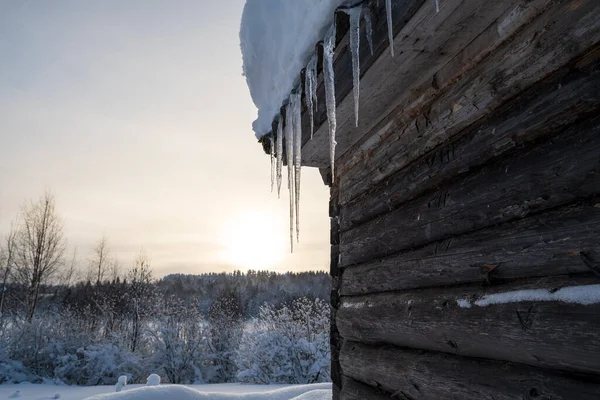 Ледяные сосульки висят на заснеженной крыше бревенчатой ванны на фоне зимнего пейзажа, в сельской местности. — стоковое фото