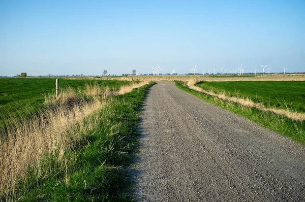 Landsvägen går genom fälten, mot bakgrund av en blå himmel och vindkraftverk, på en vårdag. Landskap. — Stockfoto