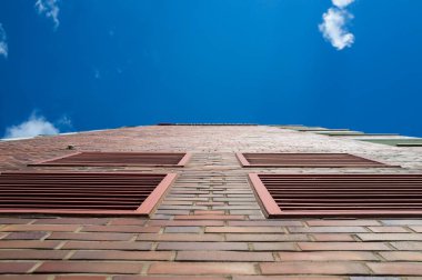 Mavi gökyüzü ve beyaz bulutların arka planında havalandırma ızgaraları olan çok katlı bir binanın tuğla duvarı. Modern şehir mimarisi. Alt görünüm.