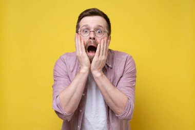 Gözlüklü adam şok olur ve korkar, ağzı açık bağırır, yanaklarından elele tutuşur ve büyük gözlerle bakar. Stüdyo, sarı arkaplan.