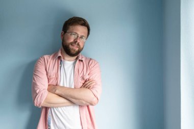 Gözlüklü, beyaz tişörtlü, pembe gömlekli, kolları göğsünde ve tatlı tatlı gülümseyen yakışıklı, sakallı bir adam. Uzayı kopyala Stüdyo, mavi arkaplan.