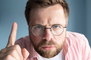 Gözlüklü kızgın sakallı adam mavi arka planda dururken işaret parmağını bir şeyi hatırlatıyormuş gibi kaldırıyor. Yakın plan..