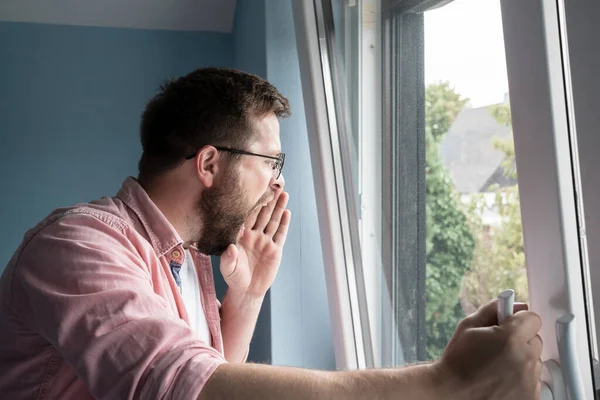 Bearded man in glasses has thrown back the sash of the window, looks into the courtyard and calls someone in a loud voice, opening mouth wide and holding hand near lips.