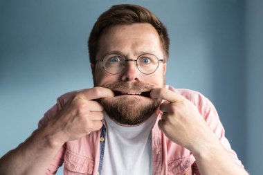 Gözlüklü tuhaf sakallı adam yüzünü buruşturdu, parmaklarını ağzına soktu, köşelere uzattı ve korkmuş gözlerle baktı. Stres ve psikolojik sağlık kavramı. Mavi arkaplan. 