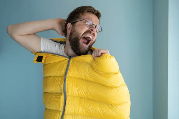 Man climbed into a yellow sleeping bag while standing indoors against a blue wall, imagining himself a tourist, yawns and stretches. Lifestyle.