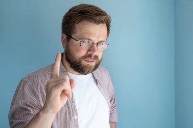 Gözlüklü sert sakallı adam işaret parmağını kaldırır, sanki bir şey hatırlatıyormuş gibi, mavi arka planda durur. Uzayı kopyala Yakın plan..