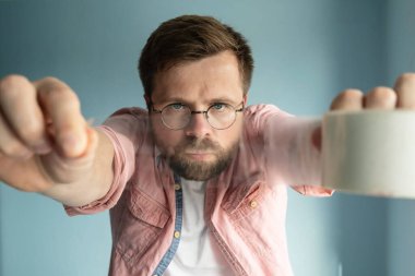 Gözlüklü ciddi sakallı bir adam uzanmış ellerinde bir rulo şeffaf yapıştırıcı bant tutuyor, odaklanmış ve bir paket hazırlamak üzere.. 