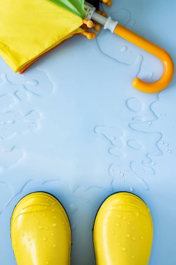 Sarı lastik su geçirmez botlar ve mavi arka plandaki su birikintisinde bir şemsiye. Yağmur mevsimi için hazırlık kavramı. Uzayı kopyala Üst Manzara. Yakın plan..