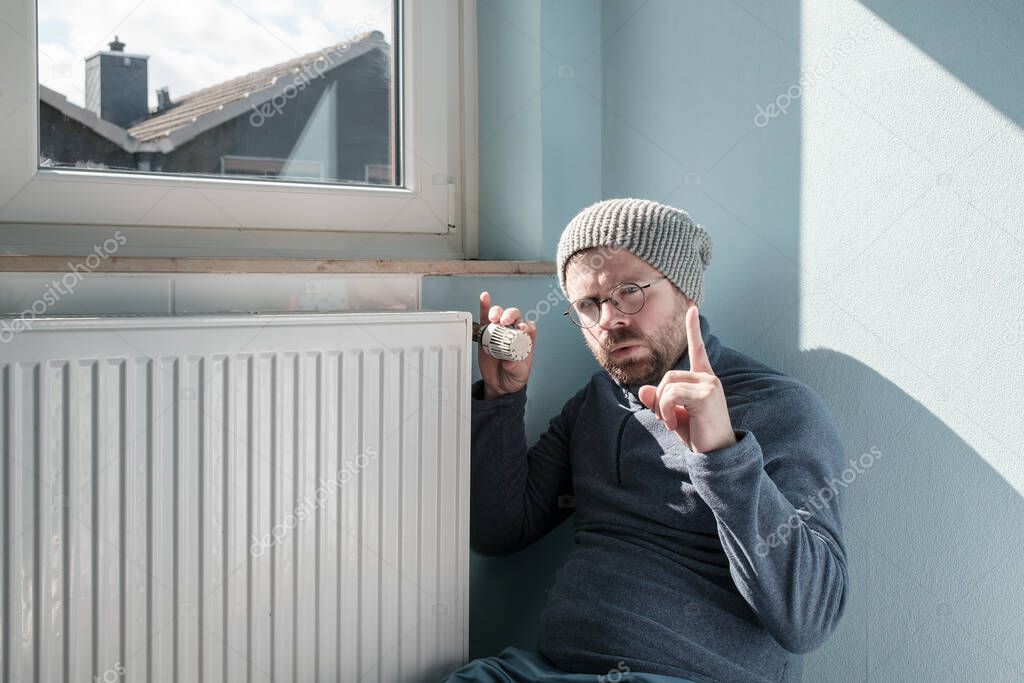 Caucasian man in a hat and a warm sweater sits near the radiator and turns the thermostat to the minimum, making a warning gesture. Concept of crisis and increase in fuel costs.
