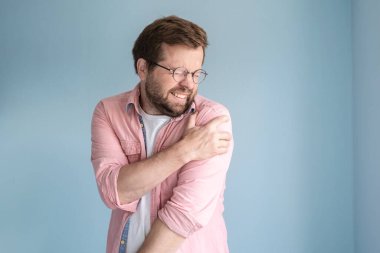 Adam omzunda şiddetli bir ağrı hissediyor, acı çekiyor, yüzünü buruşturuyor ve elini hassas noktaya koyuyor. Tıbbi konsept. İzole edilmiş, mavi arkaplan.