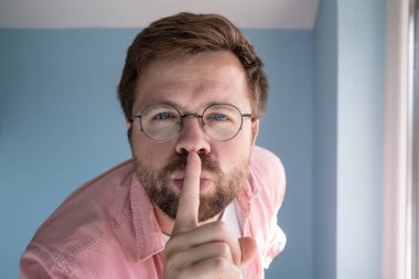 Gözlüklü beyaz bir adamın yakın plan çekiminde, işaret parmağını dudaklarına dayadı ve mavi bir duvarın arkasından sessizlik talep etti..