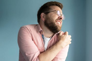 Gömlekli genç adam omzunda şiddetli bir ağrı hissediyor. Ağrıyan bölgeye masaj yapıyor, gözlerini kapatıyor ve acı çekerken dişlerini gösteriyor. Tıbbi konsept. İzole edilmiş, mavi arkaplan.