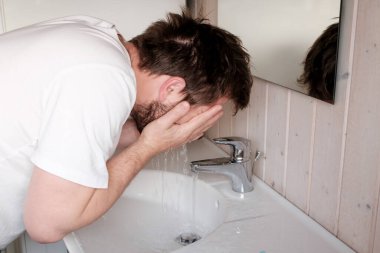 Beyaz tişörtlü bir adam yüzünü temiz akan bir su deresinin altında yıkar, sabahın erken saatlerinde, banyoda bir lavabonun üzerine eğilir. Yakın plan..