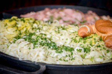 Chopped onion with herbs and chopped sausage in a large frying pan. A ruddy bun with sesame lies on top of the chopped products.