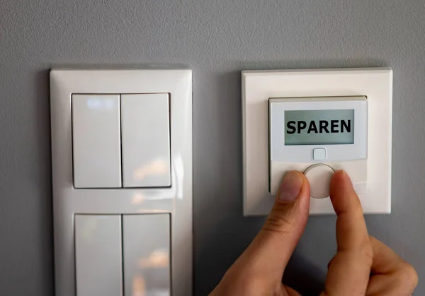 Hand changes the temperature on a electronic thermostat. The display shows the German word \'sparen\' (saving). Symbol for saving energy.