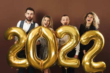 2022 yılını kutlayan bir grup güzel giyinmiş parti insanı kahverengi arka planda altın bir balonla