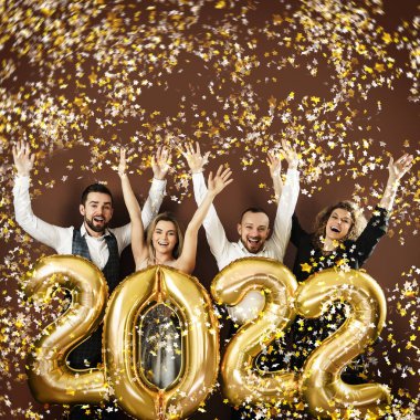 Bir grup parti insanı 2022 yılbaşını altın balonlarla ve düşen konfetilerle kutluyorlar.