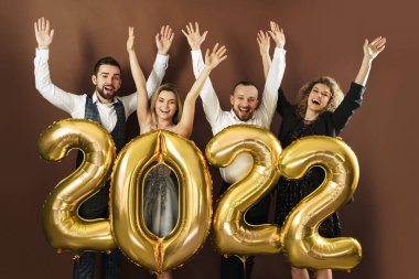 2022 yılını kutlayan bir grup güzel giyinmiş parti insanı kahverengi arka planda altın bir balonla