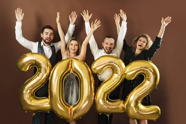 2022 yılını kutlayan bir grup güzel giyinmiş parti insanı kahverengi arka planda altın bir balonla