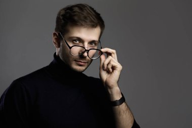 Gri arka planda balıkçı yaka ve gözlük takan genç ve zeki bir adam.