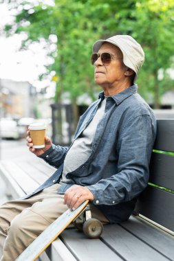 Stylish elderly man with a longboard sitting on the bench and drinking coffee in a city park clipart