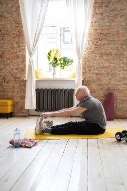 Aktif yaşlı bir adam evdeki spor yaparken sarı bir mindere oturup esniyor.