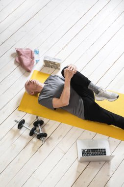 Aktif yaşlı bir adam evdeki spor yaparken sarı bir mindere uzanıyor ve esniyor.