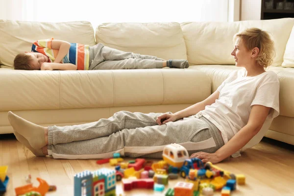 累了的男孩躺在沙发上 他的母亲玩完一个玩具后坐在地板上 — 图库照片