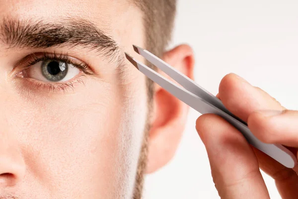 Close-up of male eye and tweezers for eyebrow grooming and shape correction