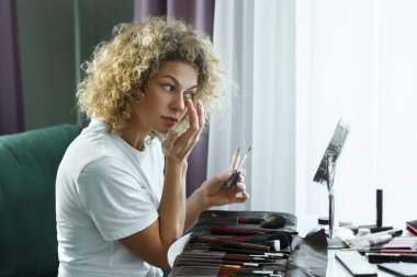 Genç ve güzel bir kadın evinde fırçayla yüzüne makyaj malzemesi sürüyor.